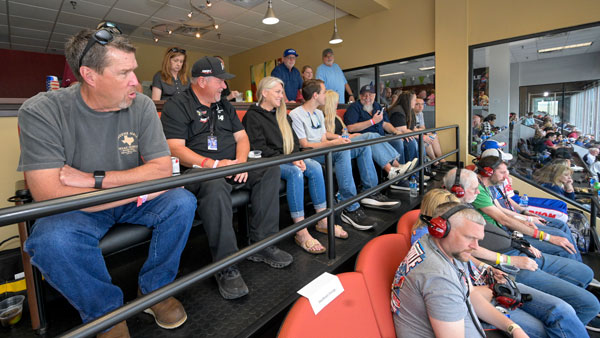 Group of people seated in an indoor viewing area at a racing event. Some are wearing headsets and race-themed clothing, while others sit casually in chairs or on a raised platform. Large windows in the background provide a view of the racetrack and spectators outside.