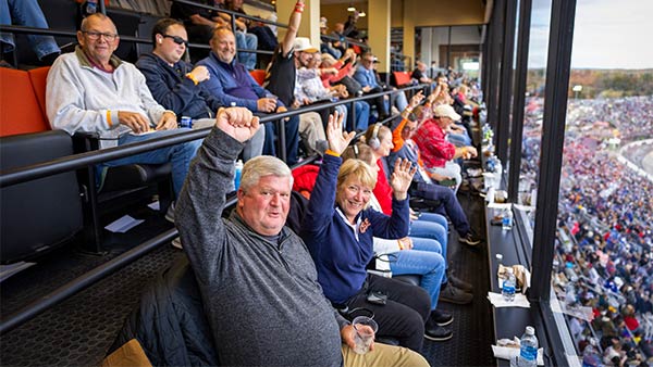 Fans seated in a covered grandstand suite cheer and raise their hands while watching the race through large windows at Martinsville Speedway.