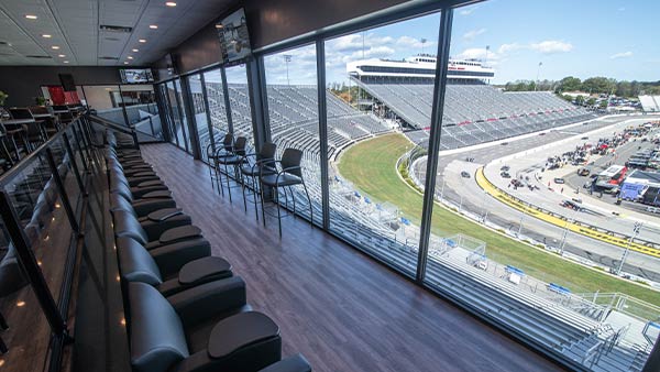 Interior view of Club Panorama suite at Martinsville Speedway, showing rows of black leather seats and barstools facing large floor-to-ceiling windows that overlook the racetrack and grandstands on a clear day.