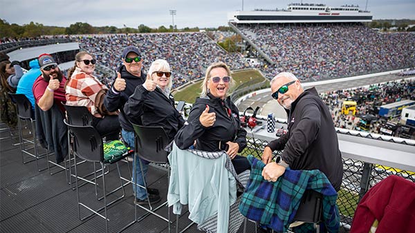 A group of fans smiles and gives thumbs up while seated on high chairs along the SkyDeck railing overlooking the packed Martinsville Speedway.
