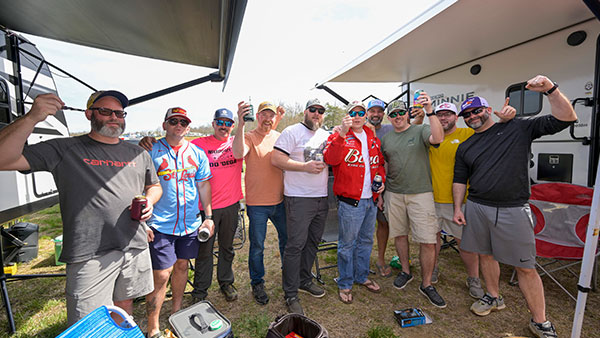 A group of approximately ten adult men stand closely together outdoors between two large RVs in a campground setting. A shade canopy extends overhead on the left side of the image. The men are dressed casually in T-shirts, shorts, baseball caps, and sunglasses, with several wearing racing-themed apparel. Many are holding canned beverages and raising them toward the camera in a celebratory gesture. Coolers and camping supplies are visible near their feet on the grass. The group appears cheerful and relaxed, suggesting a friendly tailgating atmosphere during a race weekend.