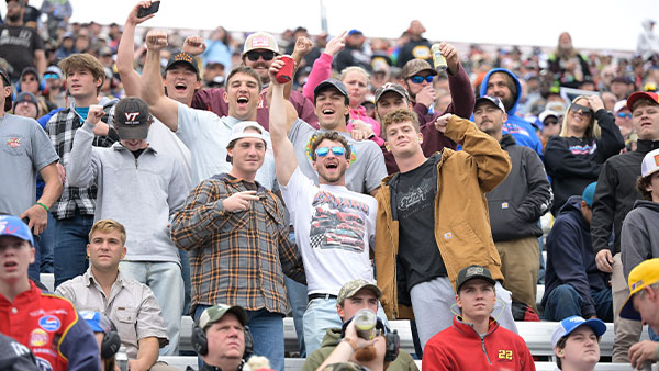 A large crowd of fans fills a section of racetrack grandstands. In the foreground, a group of young adult men stand and cheer with raised fists and open mouths, expressing excitement. They wear casual clothing including hoodies, flannel shirts, T-shirts, and baseball caps, some featuring racing or collegiate logos. Other spectators sit and stand behind them, also watching the event. The atmosphere appears energetic and celebratory, consistent with a live race moment at a NASCAR event.