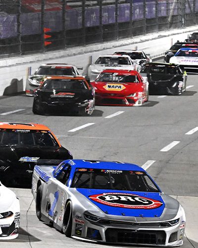 Two late model stock cars race side-by-side on a paved straightaway under bright track lighting. The car on the left is primarily white with black accents and “Pembelton Realty” displayed across the windshield banner. The car on the right is silver with a blue hood featuring a large red circular logo. Several additional race cars follow closely behind in staggered formation. The concrete retaining wall lines the left side of the track, marked with vertical colored stripes, while dashed white lane markings are visible on the racing surface. The scene conveys close competition at speed during a nighttime event.