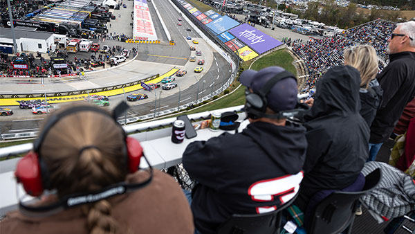 A group of spectators sits in elevated outdoor seating overlooking a paved oval racetrack during a NASCAR event. Several people in the foreground wear headsets and jackets, seated at a counter-style ledge with drinks placed in front of them. Below, multiple brightly colored stock cars race through a left-hand turn. The infield is visible with haulers, pit road activity, and sponsor signage painted on the track surface. Grandstands across the track are filled with fans.