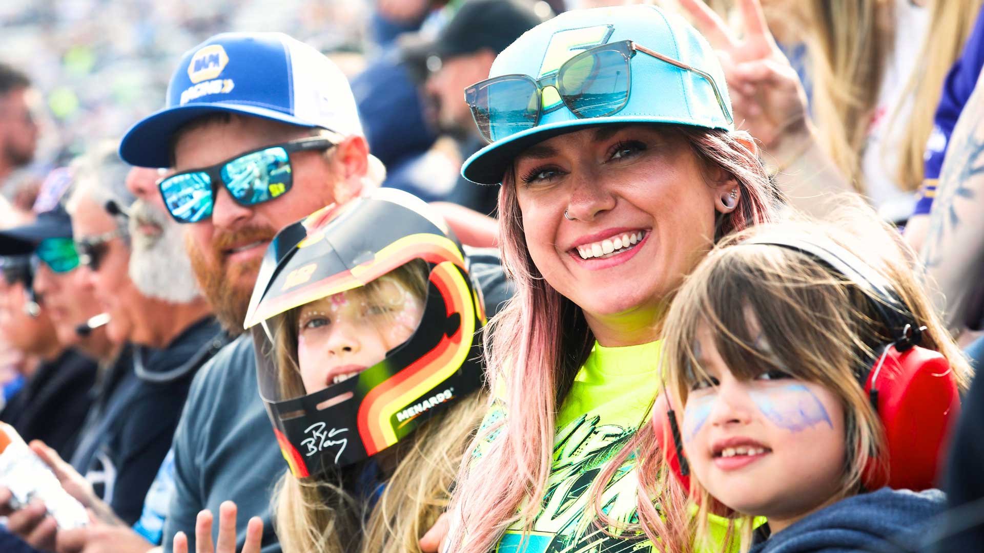 Family wearing racing gear and face paint smiling in the grandstands at a NASCAR race, with one child wearing a driver helmet and another wearing hearing protection headphones.